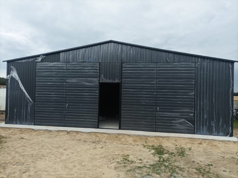 Nowy, grafitowy garaż blaszany z dwoma parami wrót, częściowo zabezpieczony folią ochronną, na betonowej wylewce, na tle pochmurnego nieba.