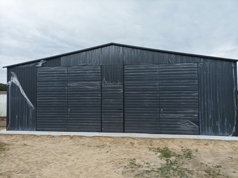 Nowy, grafitowy garaż blaszany z dwoma bramami rolowanymi, częściowo zabezpieczony folią ochronną, na betonowej wylewce, z widocznym piaskowym podłożem.