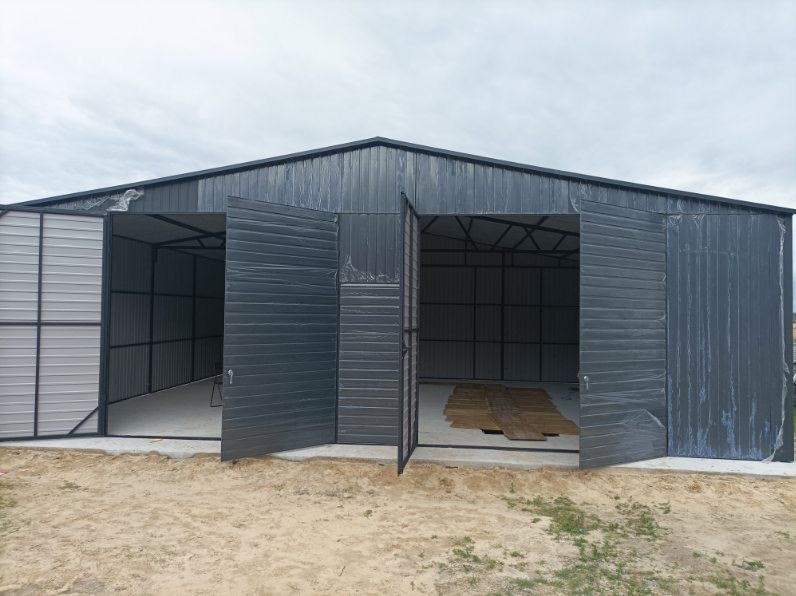 Szary garaż blaszany z otwartymi wrotami, widoczne wnętrze z betonową podłogą i drewnianymi deskami. Garaż usytuowany na piaszczystym podłożu pod pochmurnym niebem.