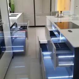 Nowoczesna kuchnia z otwartymi szufladami podświetlonymi LED, widoczne sprzęty AGD i wyspa kuchenna z blatem. W tle osoba w salonie.