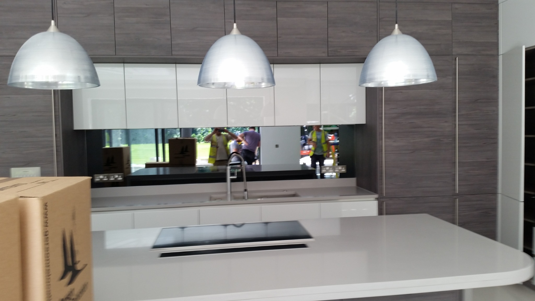 Nowoczesna kuchnia z białymi szafkami i szarymi panelami drewnopodobnymi. Lustrzany panel nad blatem odbija otoczenie. Trzy srebrne lampy wiszą nad wyspą.