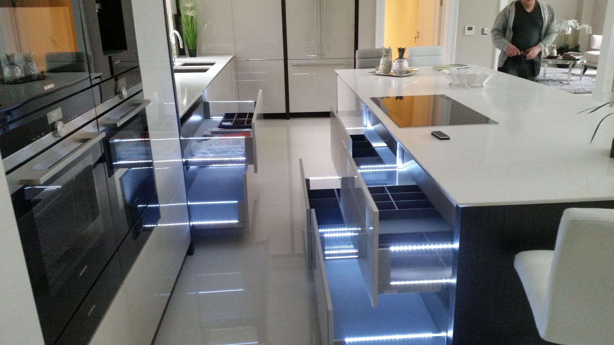 Nowoczesna kuchnia z otwartymi szufladami podświetlonymi LED, widoczne sprzęty AGD i wyspa kuchenna z blatem. W tle osoba w salonie.