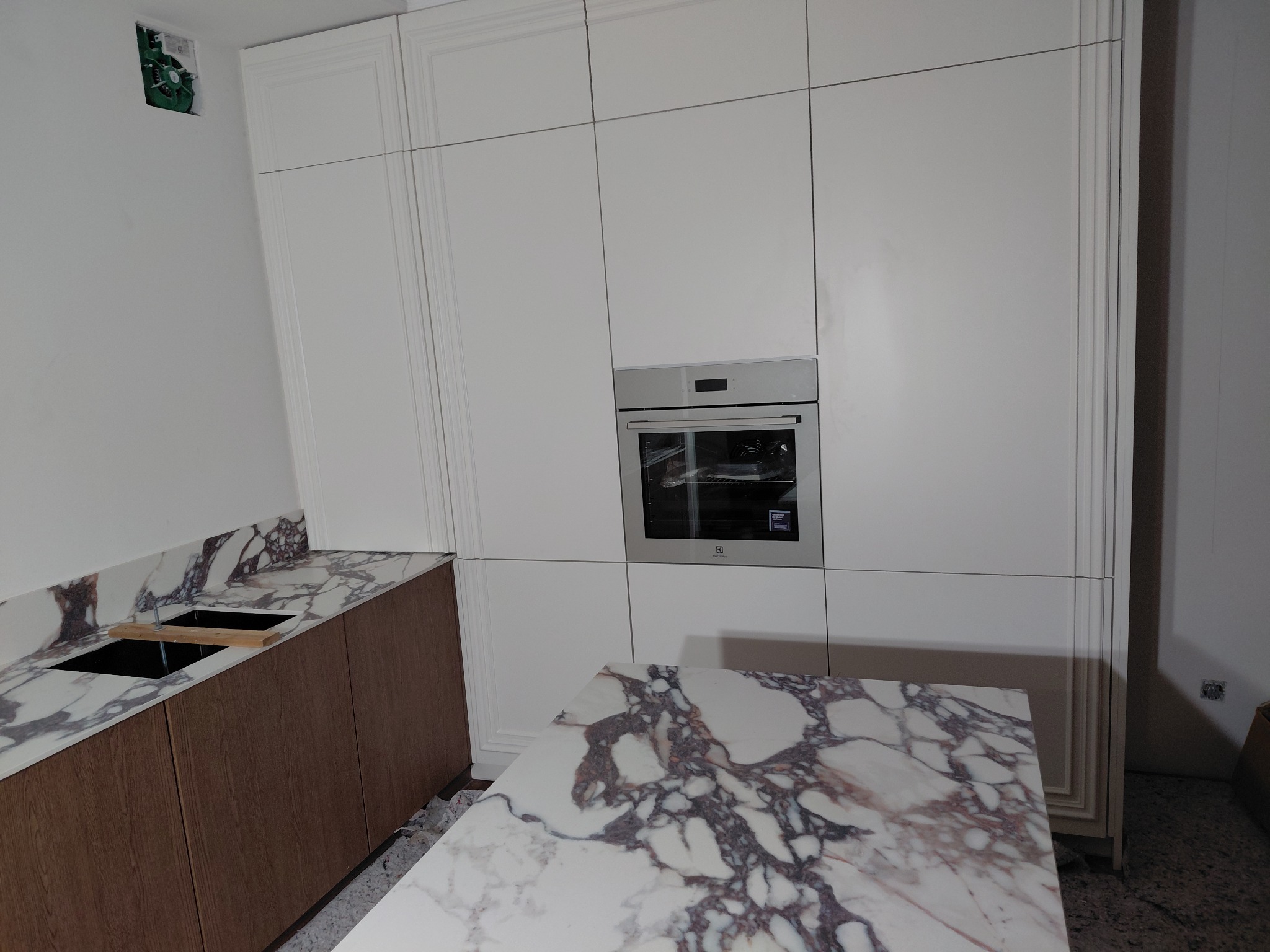Nowoczesna kuchnia z białymi szafkami i blatem z marmuru. Widoczny piekarnik w zabudowie i fragment niezabudowanej ściany z instalacją elektryczną.