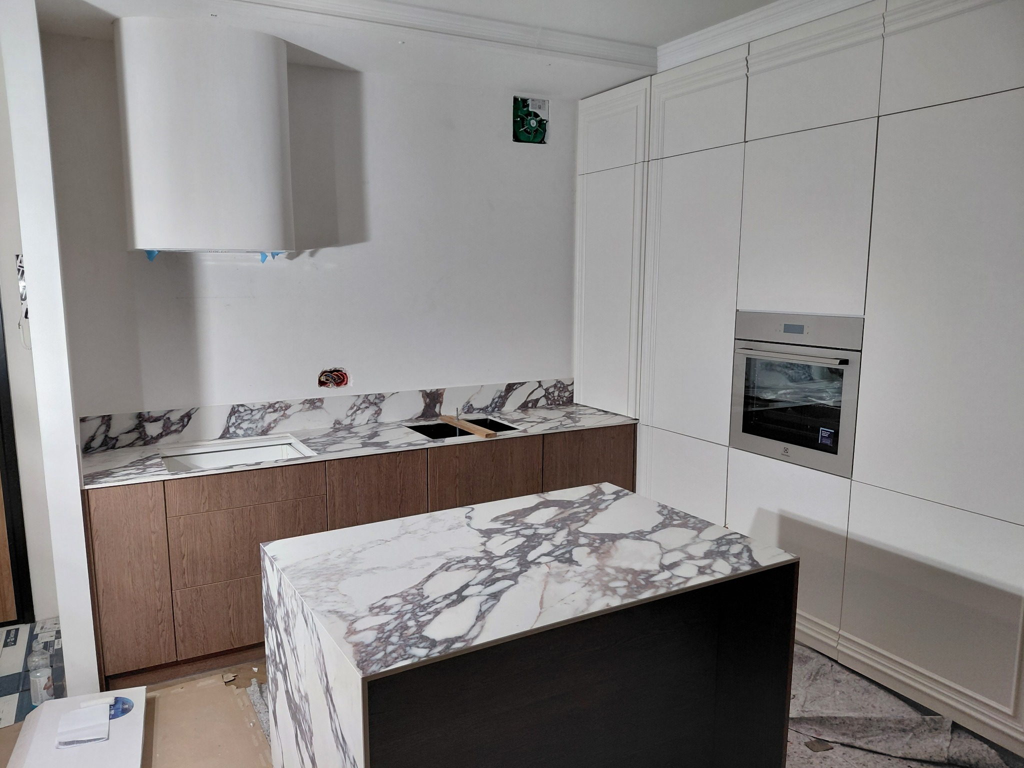 Nowoczesna kuchnia z wyspą i marmurowym blatem. Białe szafki górne z piekarnikiem, brązowe szafki dolne. Na ścianie biały okap. Podłoga zabezpieczona folią.