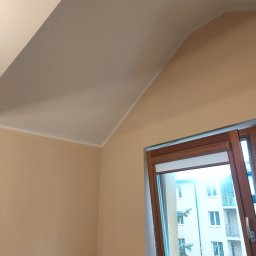 Kompleksowe Wykończenia Wnętrz Marek Biskowski - Beżowe ściany i biały sufit na poddaszu po malowaniu, widok na otwarte okno z drewnianą ramą i budynek za oknem. Świeżo pomalowane wnętrze.