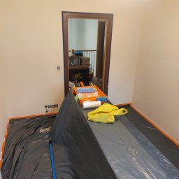 Kompleksowe Wykończenia Wnętrz Marek Biskowski - Przygotowania do malowania: zabezpieczona folią podłoga, taśma malarska na ścianach i futrynach, kuweta z wałkiem i pędzle. Widok na wnętrze domu przez otwarte drzwi.
