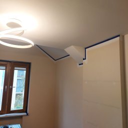 Kompleksowe Wykończenia Wnętrz Marek Biskowski - Przygotowanie do malowania ścian w Świętajno: pokój zabezpieczony folią, taśma malarska na suficie i oknach, gotowy do nałożenia farby. Jasne wnętrze z nowoczesnym oświetleniem.