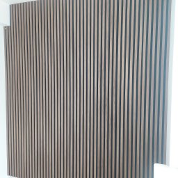 Kompleksowe Wykończenia Wnętrz Marek Biskowski - Ściana wykończona panelami lamelowymi w odcieniach brązu, tworzącymi pionowy wzór. Widoczny fragment podłogi i narzędzie pomiarowe w lewym dolnym rogu.