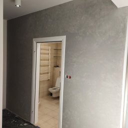 Kompleksowe Wykończenia Wnętrz Marek Biskowski - Ściana w łazience po nałożeniu dekoracyjnej, szarej gładzi weneckiej. Widoczne gniazdka elektryczne i fragment łazienki z grzejnikiem drabinkowym przez otwarte drzwi.