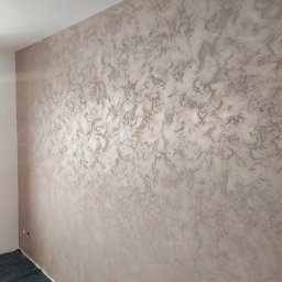 Kompleksowe Wykończenia Wnętrz Marek Biskowski - Ściana w pokoju po nałożeniu dekoracyjnej, beżowo-srebrnej gładzi. Widoczne zabezpieczenie podłogi folią malarską i fragment ściany bocznej. Efektowna faktura na ścianie.