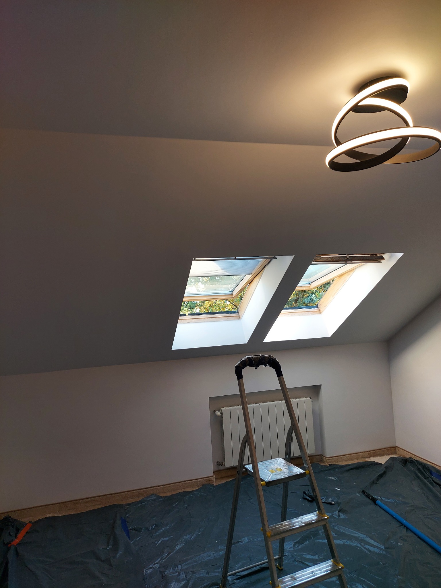 Poddasze po remoncie: świeżo pomalowane ściany, okna dachowe, nowoczesna lampa. Rozłożona folia ochronna i drabina sugerują prace wykończeniowe.