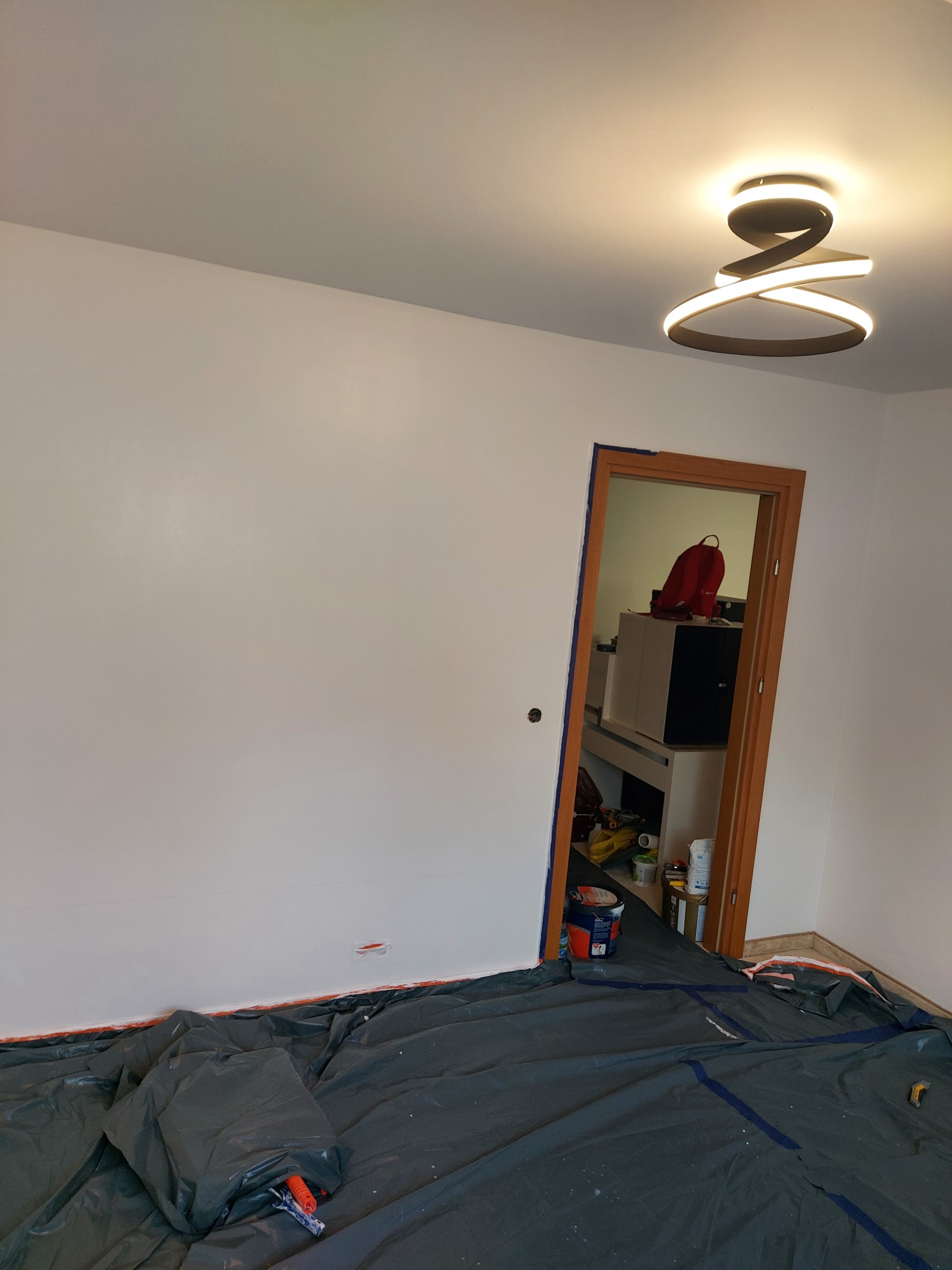 Świeżo pomalowane, białe ściany pokoju z widocznym zabezpieczeniem podłogi folią malarską i nowoczesną lampą sufitową. Widoczne drzwi do kolejnego pomieszczenia.