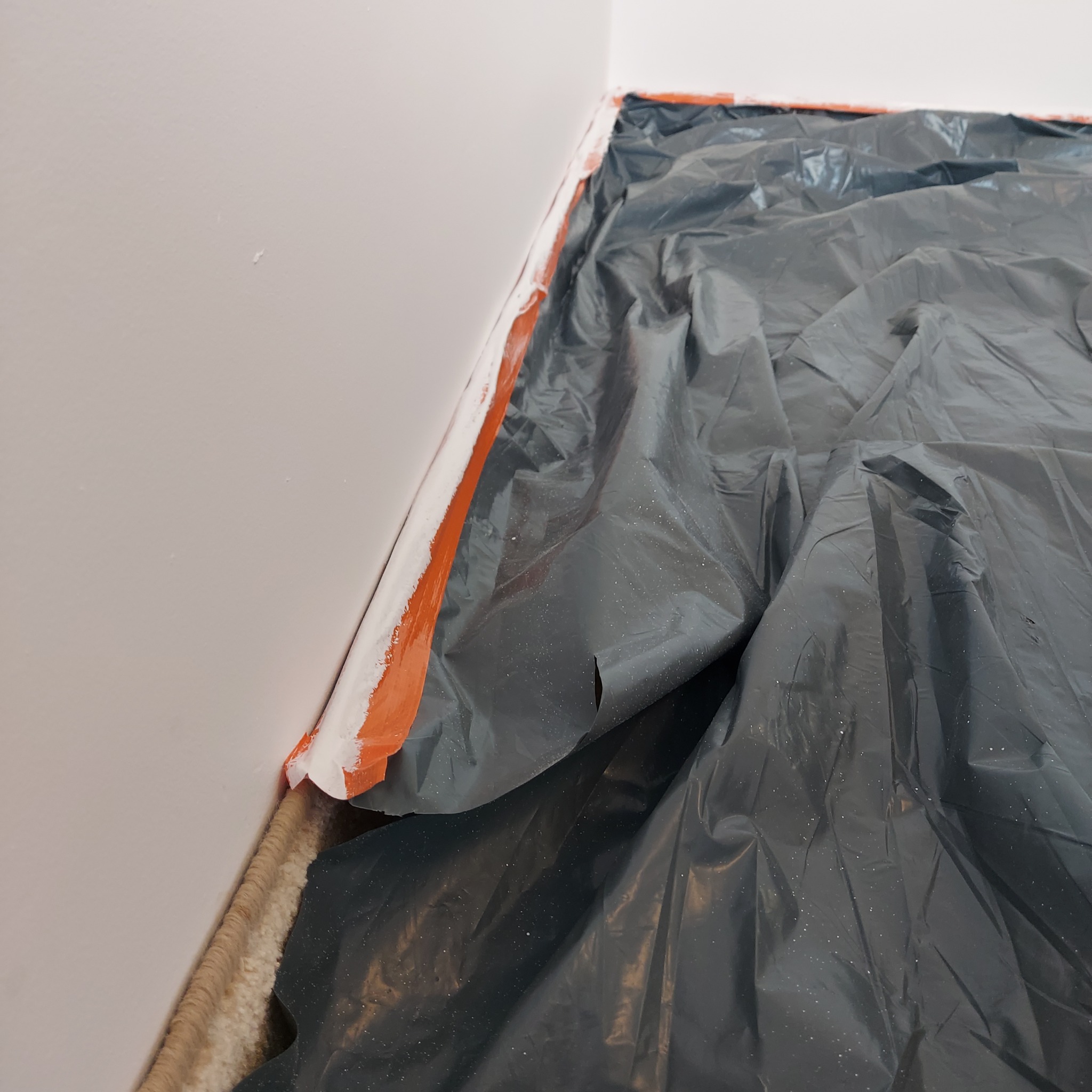 Przygotowanie do malowania: zabezpieczona folią podłoga i taśmą malarską ściana w rogu pokoju. Widoczne świeżo pomalowane fragmenty ściany.