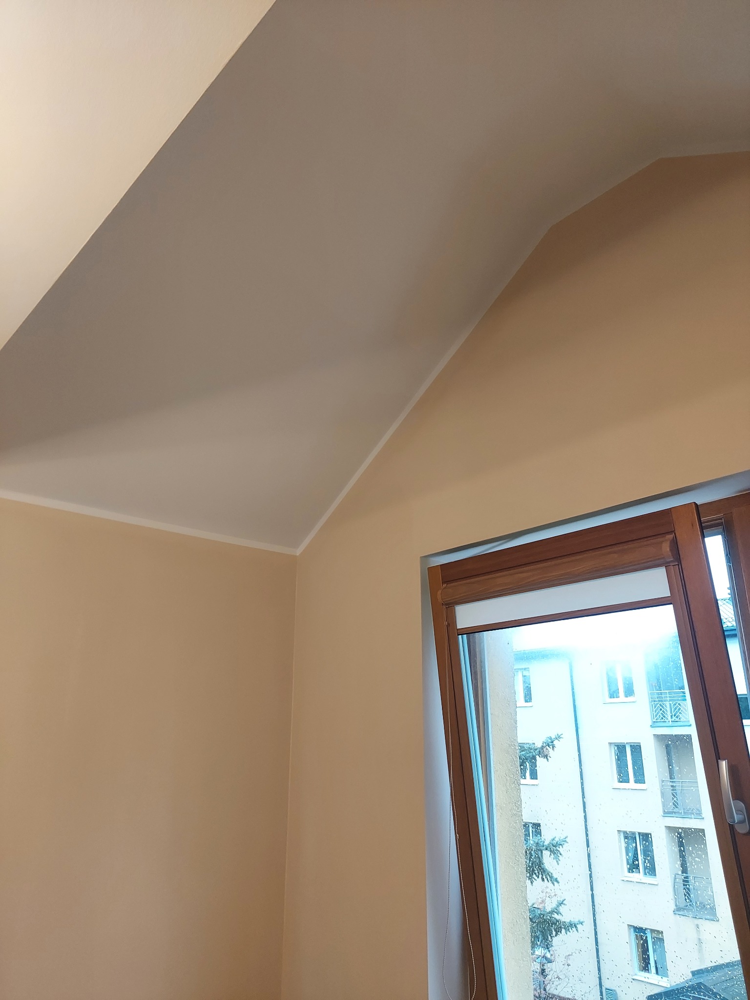 Beżowe ściany i biały sufit na poddaszu po malowaniu, widok na otwarte okno z drewnianą ramą i budynek za oknem. Świeżo pomalowane wnętrze.