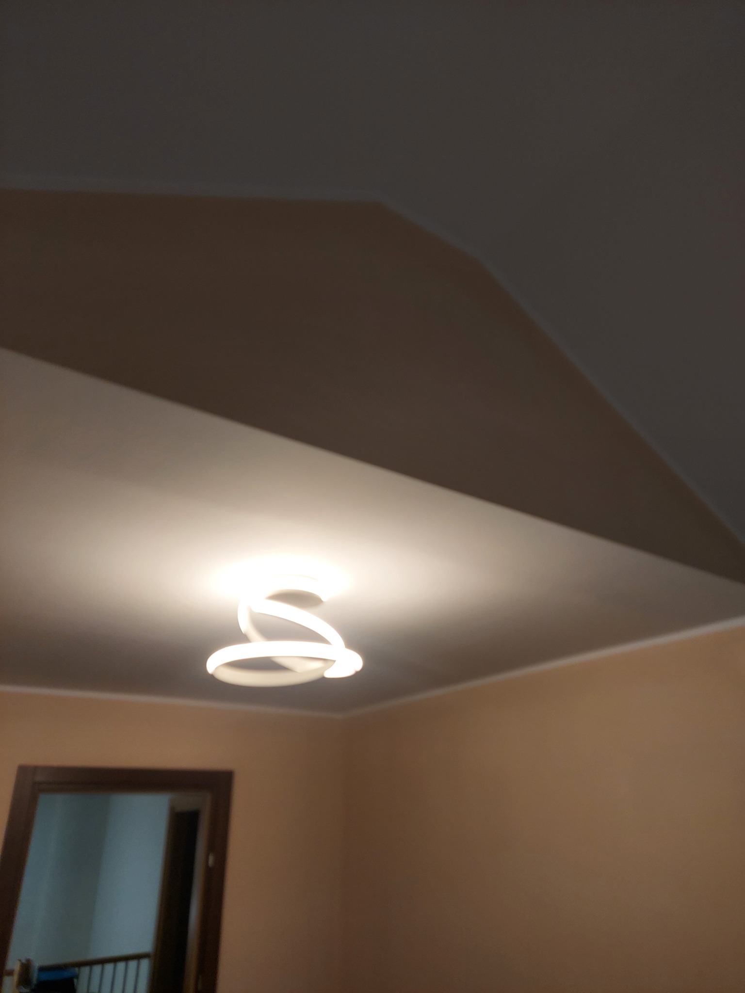 Beżowe wnętrze z nowoczesną lampą sufitową. Widoczny fragment skośnego sufitu i drzwi w tle. Ściany w ciepłym odcieniu beżu.