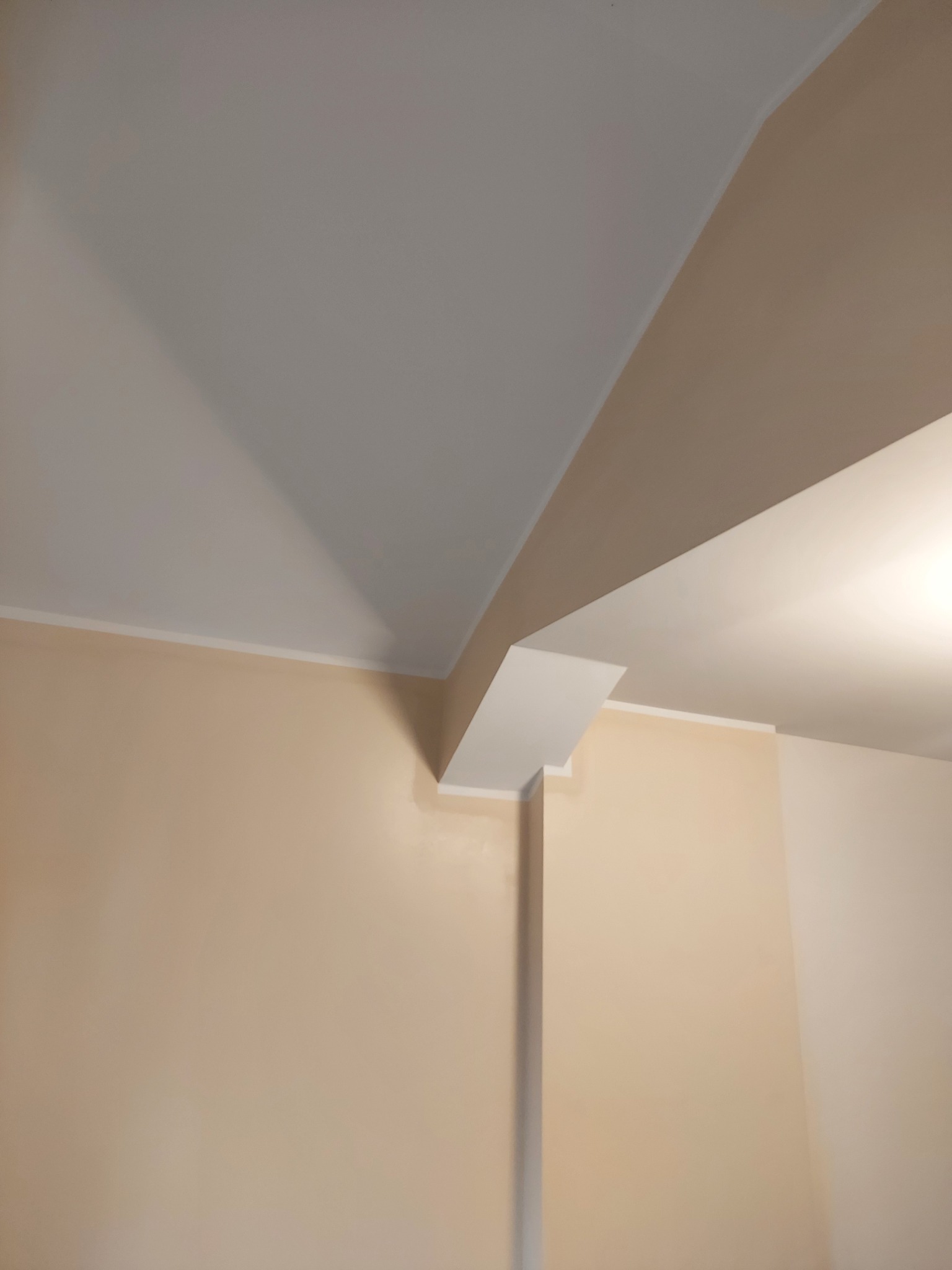 Jasne wnętrze po remoncie: kremowa ściana, biały sufit z wystającym elementem konstrukcyjnym. Widoczne listwy przypodłogowe i sufitowe. Ujęcie z dołu, podkreślające geometrię.