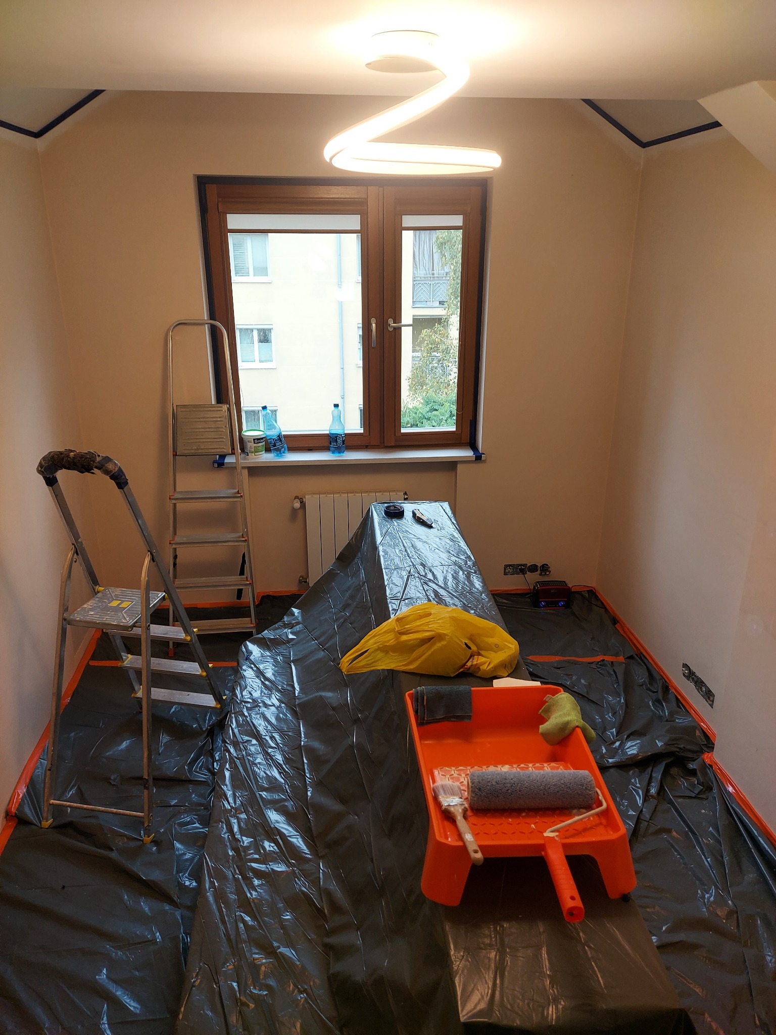 Pokój przygotowany do malowania: folia ochronna, drabina, kuweta z wałkiem i pędzlem. Ściany w jasnym odcieniu, okno z widokiem na budynek. Nowoczesna lampa sufitowa.
