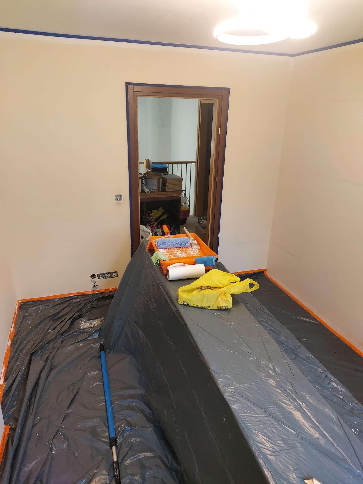 Przygotowania do malowania: zabezpieczona folią podłoga, taśma malarska na ścianach i futrynach, kuweta z wałkiem i pędzle. Widok na wnętrze domu przez otwarte drzwi.