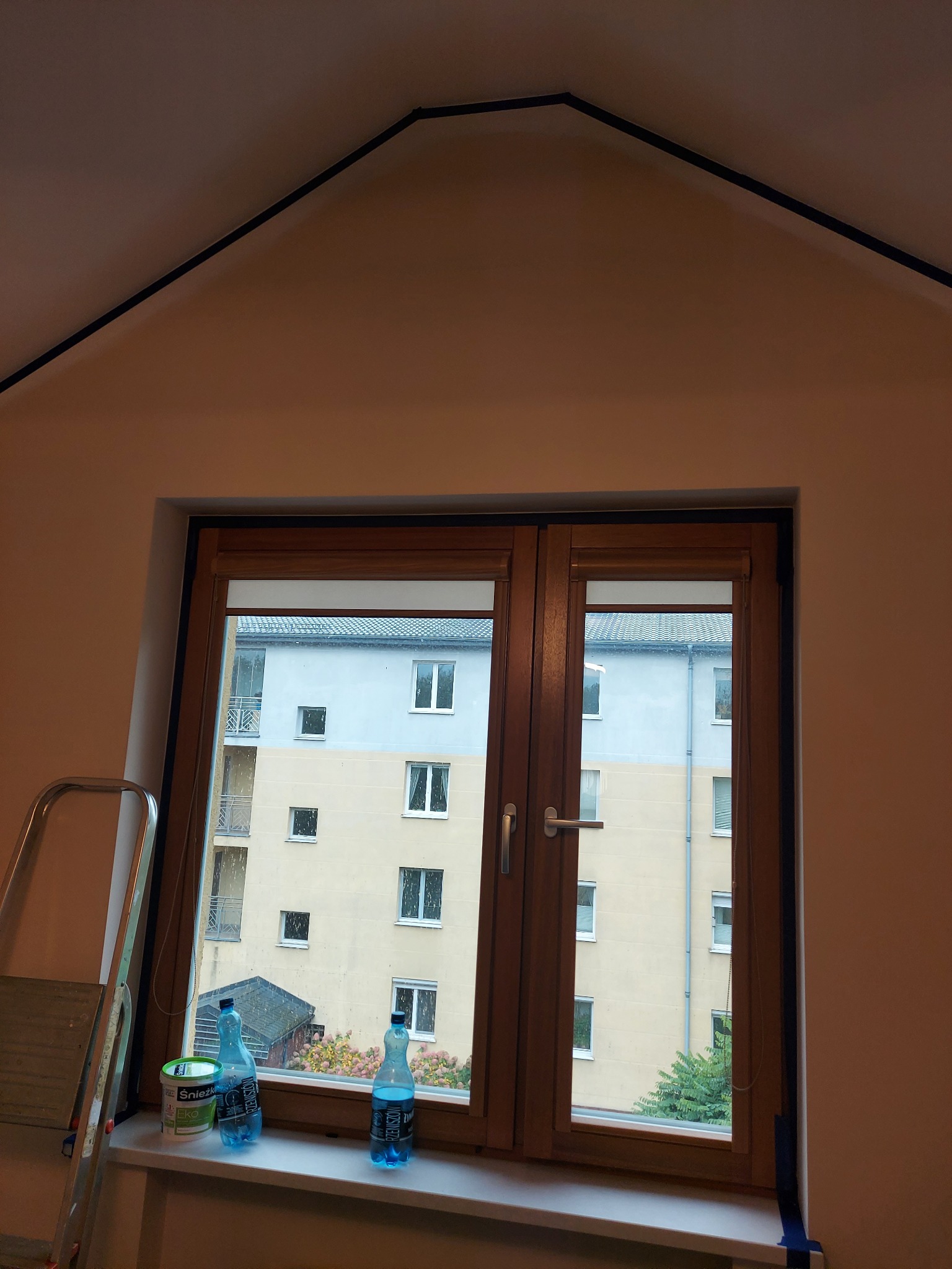 Ściana po malowaniu w pokoju na poddaszu, widok na okno z farbą i drabiną. Świeżo pomalowane ściany w odcieniach beżu, czarne listwy przy suficie.