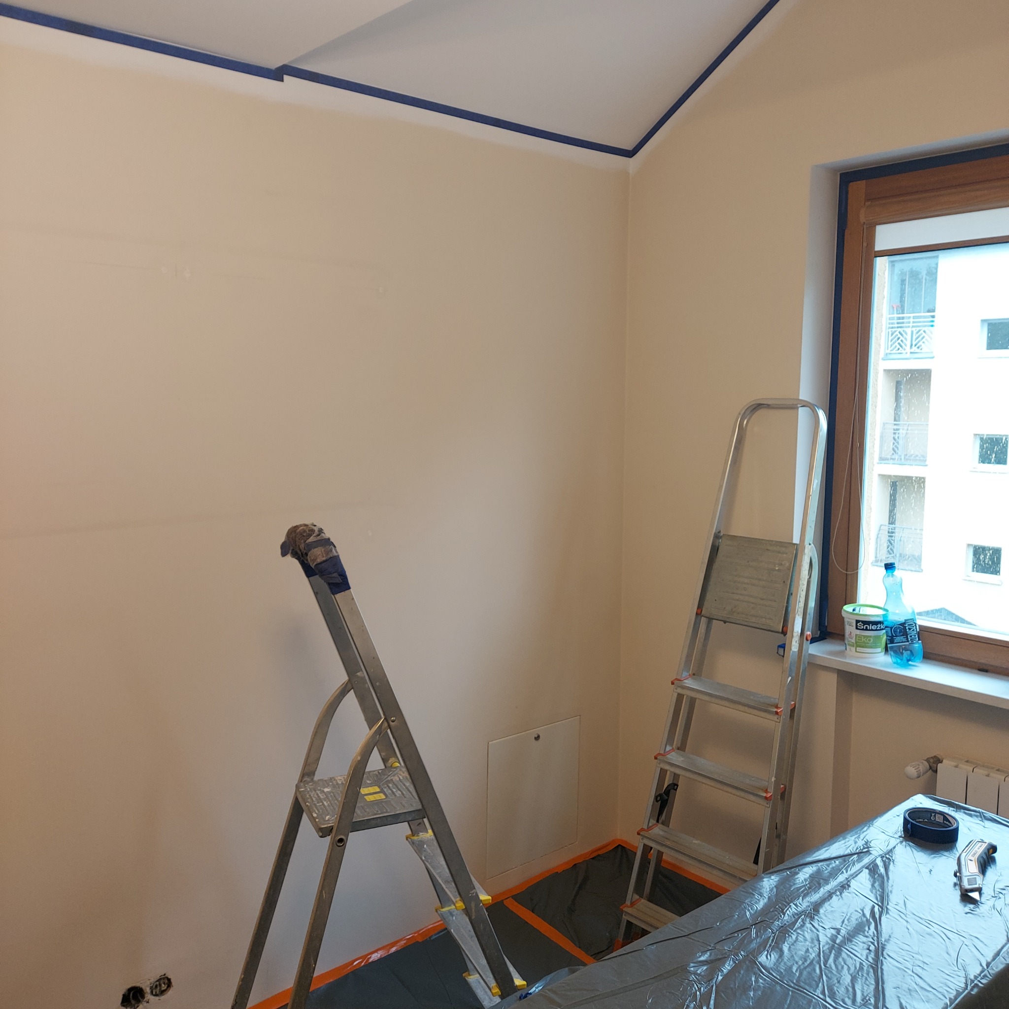 Pokój przygotowany do malowania: zabezpieczone ściany taśmą, podłoga folią, drabiny ustawione przy ścianach. Widoczne puszki z farbą na parapecie.