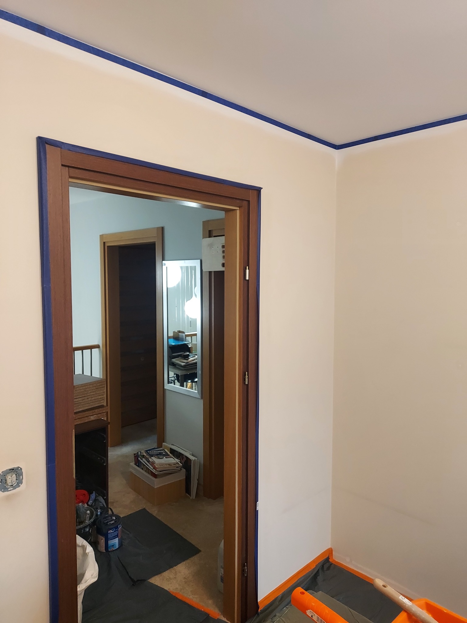 Przygotowanie do malowania: ściany i futryny zabezpieczone niebieską taśmą malarską, podłoga przykryta folią ochronną. Widoczne narzędzia i farby.