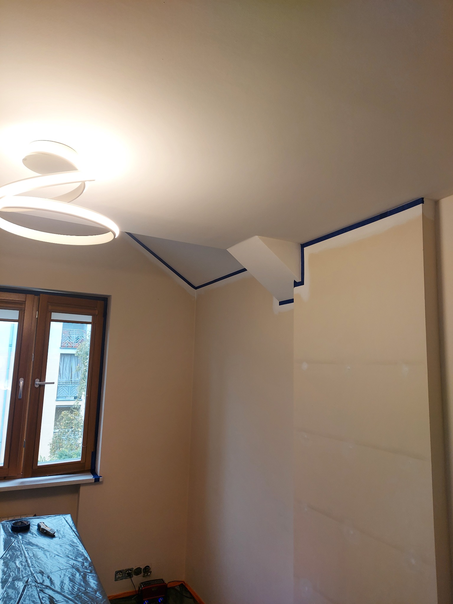 Przygotowanie do malowania ścian w Świętajno: pokój zabezpieczony folią, taśma malarska na suficie i oknach, gotowy do nałożenia farby. Jasne wnętrze z nowoczesnym oświetleniem.