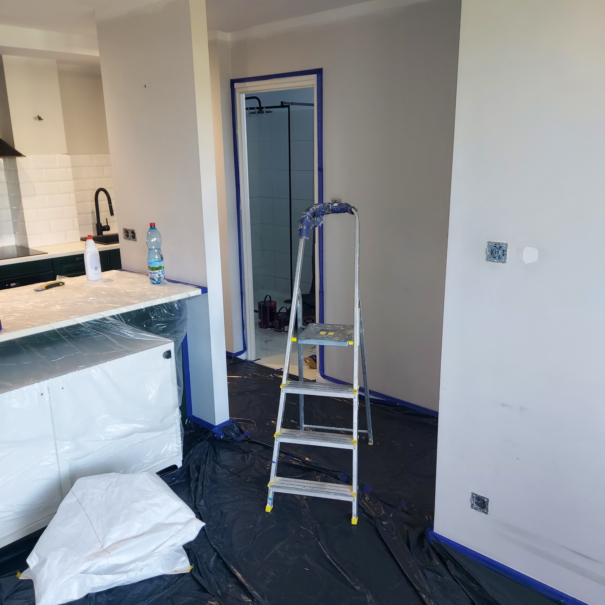 Wnętrze w trakcie malowania: zabezpieczona folią kuchnia, drabina przy ścianie z taśmą malarską. Widoczne puszki farb i niedokończone gniazdka.
