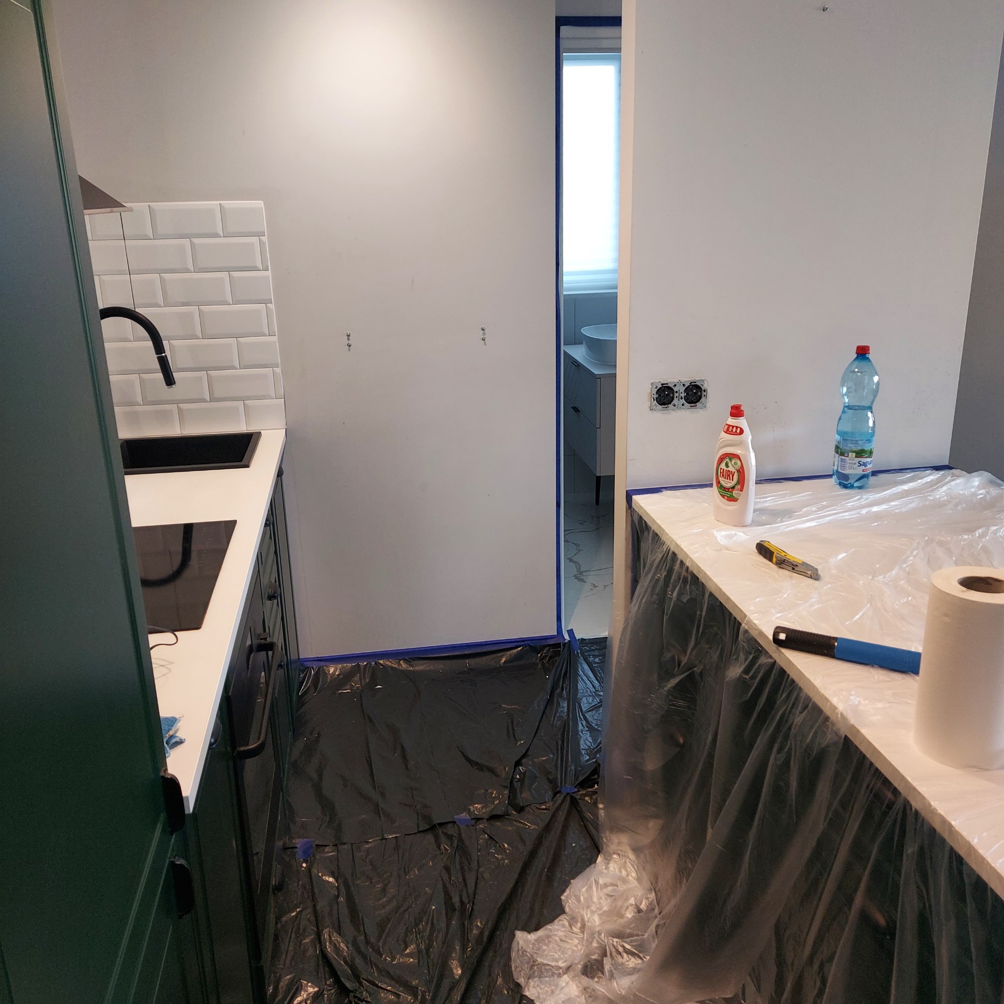 Przygotowania do malowania: zabezpieczona folią i taśmą kuchnia z widocznymi narzędziami malarskimi i środkami czystości. Widok na łazienkę w tle.