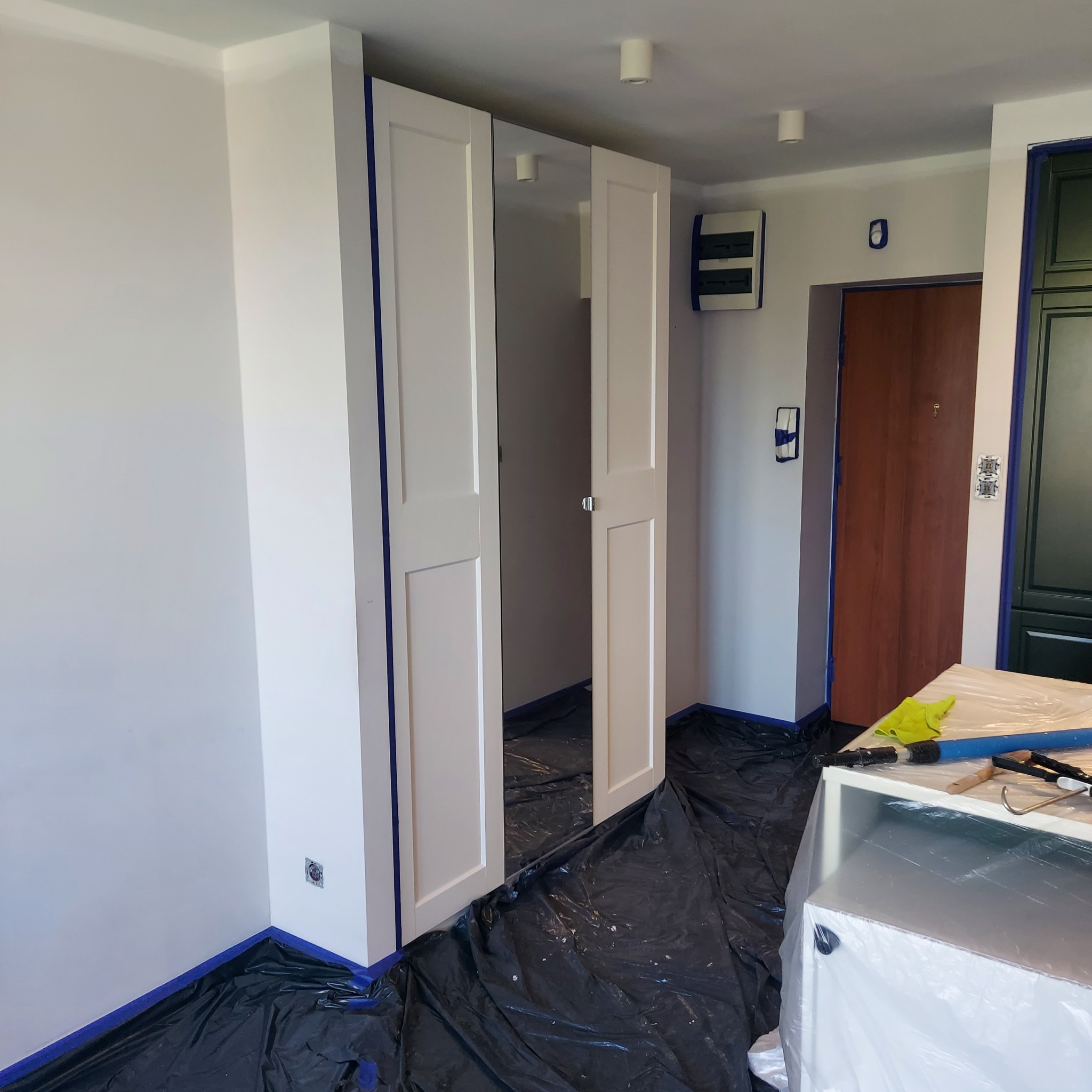 Biała szafa z lustrami zabezpieczona taśmą malarską i folią, przygotowana do malowania ścian. Widoczne narzędzia malarskie i zabezpieczone podłoże.