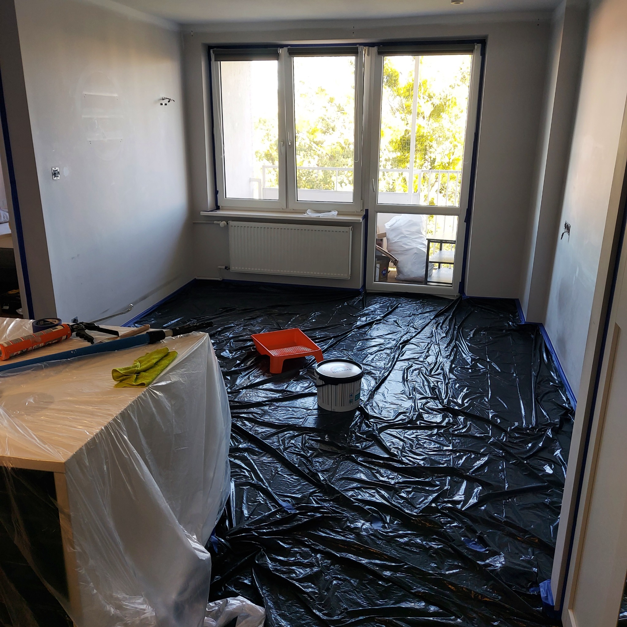 Pokój przygotowany do malowania: podłoga zabezpieczona folią, ściany z taśmą malarską, widoczny kuweta i pędzel. Meble zakryte folią ochronną. Jasne wnętrze z oknem.
