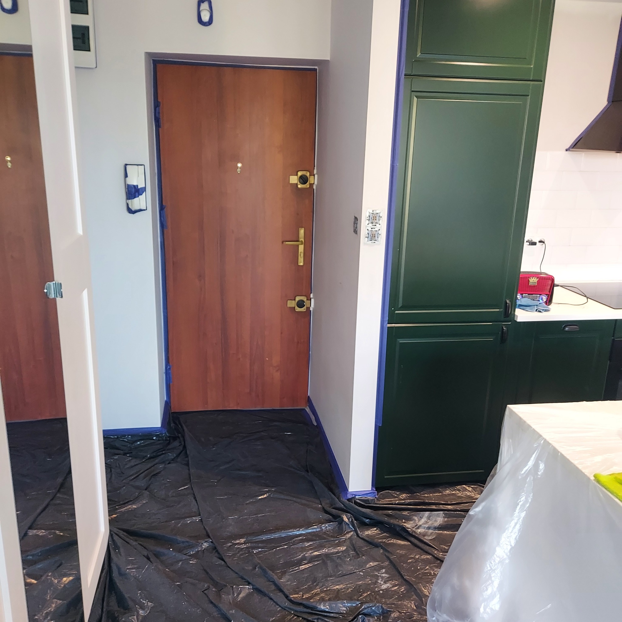 Przygotowanie do malowania: zabezpieczona folią podłoga, taśmą oklejone futryny drzwi i szafki kuchenne. Widoczny fragment kuchni i przedpokoju.