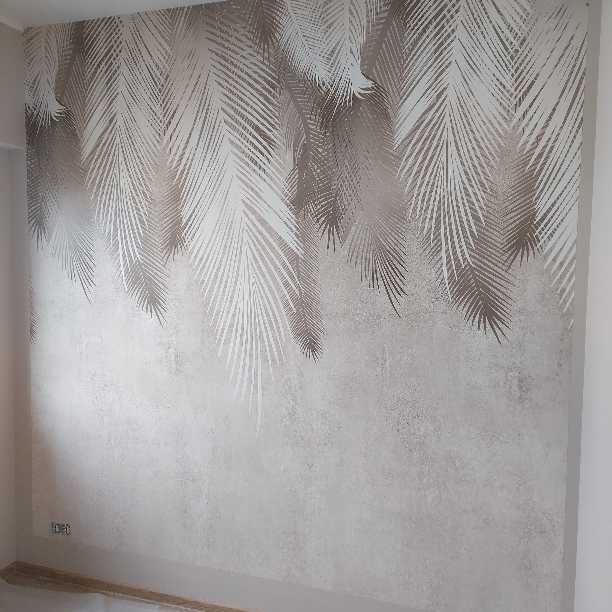 Ściana z dekoracyjnym wzorem liści palmowych w odcieniach beżu i bieli. Widoczne gniazdka elektryczne i zabezpieczona folią podłoga.