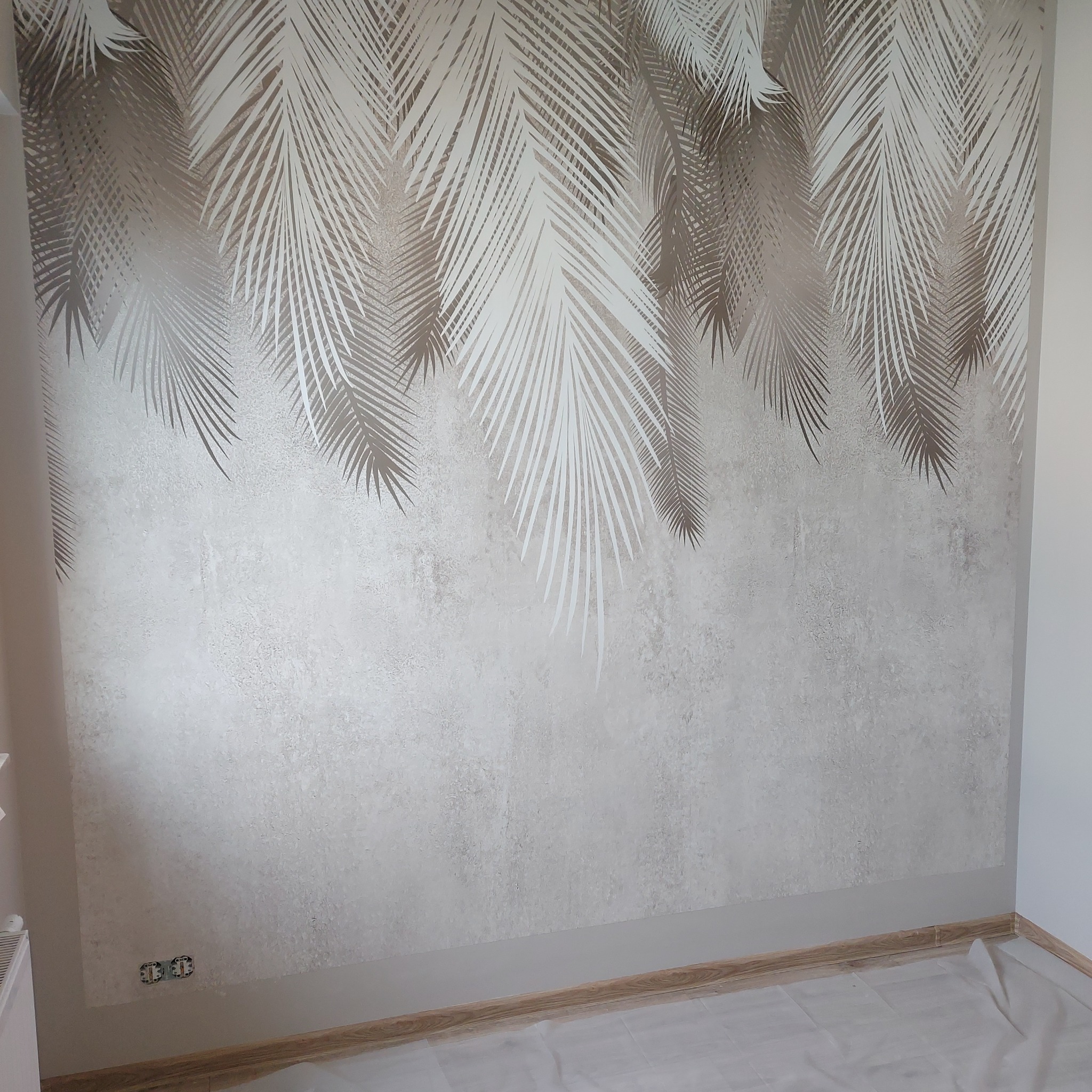 Ściana po malowaniu z dekoracyjnym wzorem liści palmowych w odcieniach beżu i bieli. Widoczne gniazdka elektryczne i zabezpieczona folią podłoga.