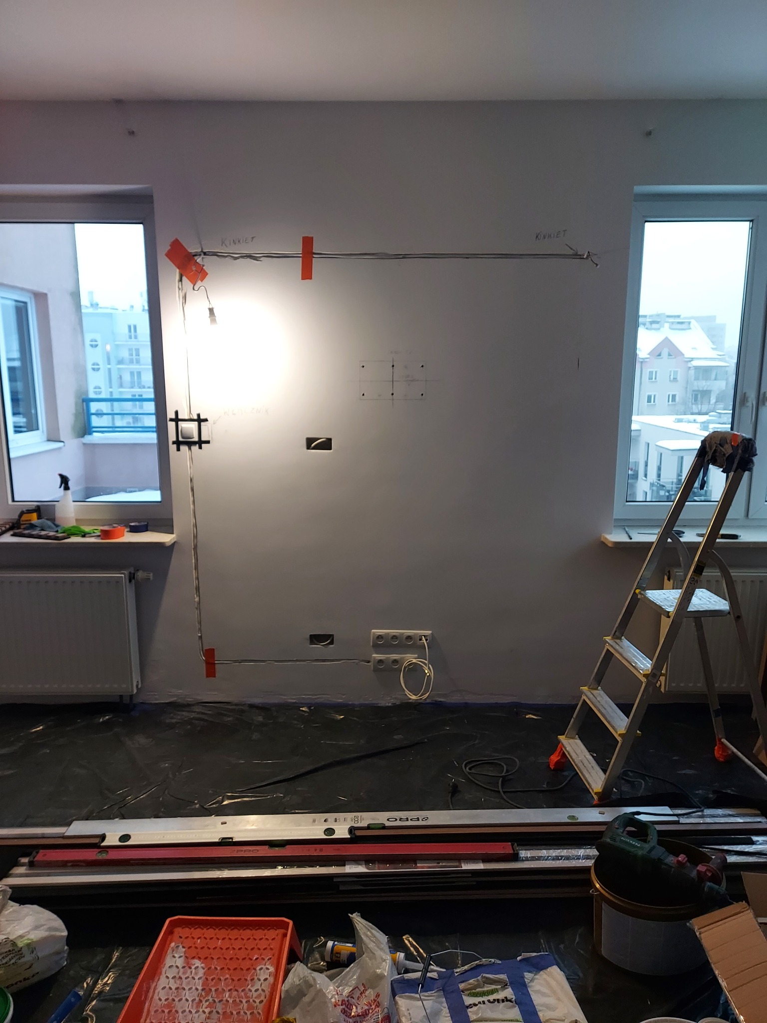 Ściana w trakcie przygotowania do montażu oświetlenia: widoczne kable, oznaczenia, puszki elektryczne, drabina i narzędzia na podłodze zabezpieczonej folią.