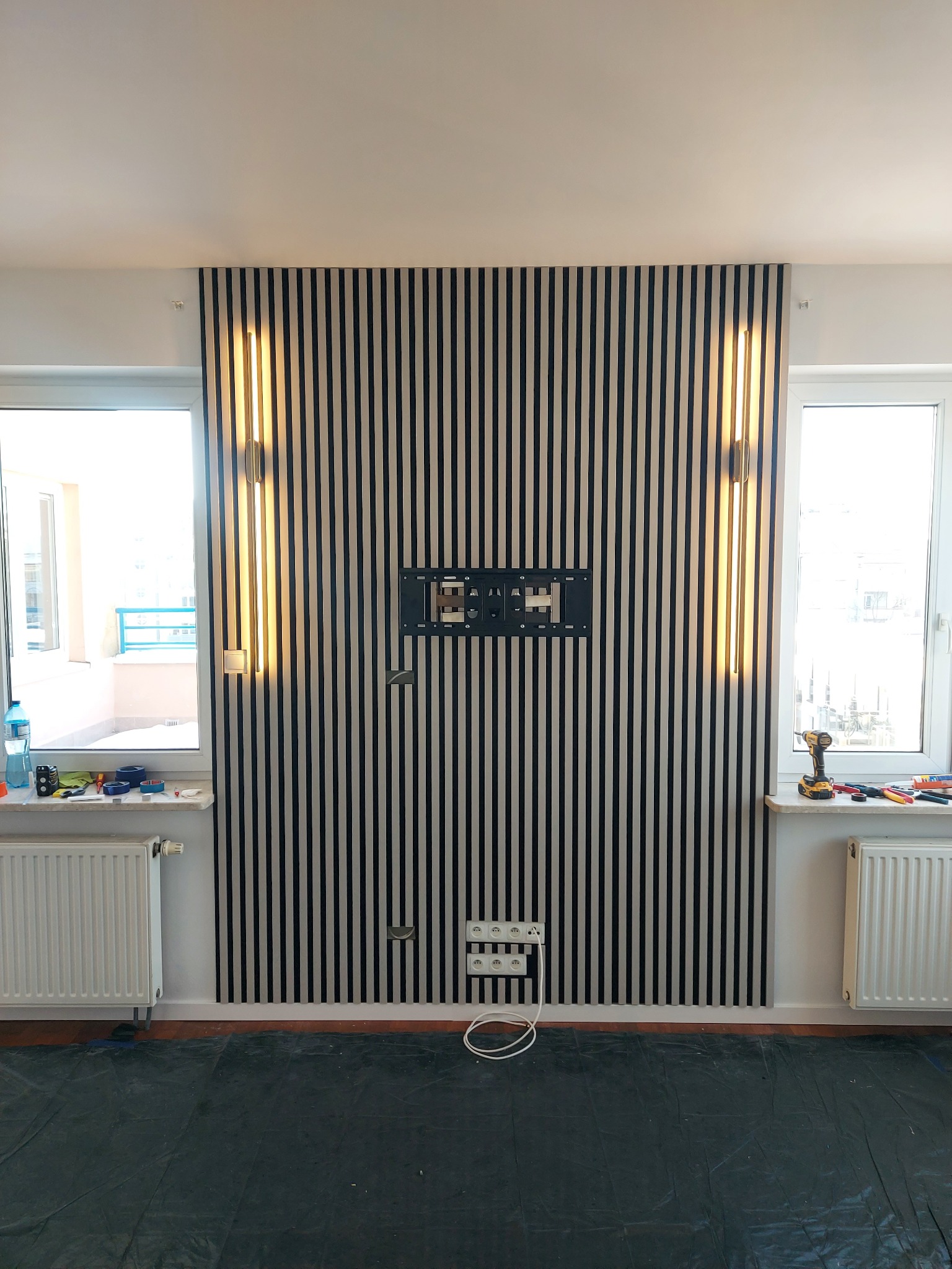 Ściana z lameli w czarno-białe pasy, oświetlenie boczne, gniazdka i uchwyt TV. Wykończenie wnętrza w Świętajno, widoczne narzędzia i folia ochronna na podłodze.