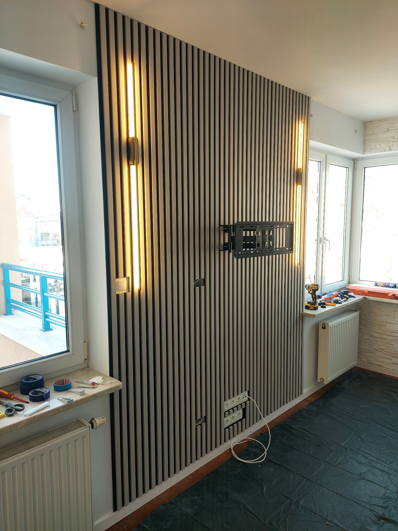 Ściana z drewnianych lameli w odcieniach szarości, oświetlona pionowymi lampami. Widoczny uchwyt na telewizor, narzędzia na parapecie i zabezpieczona folią podłoga.
