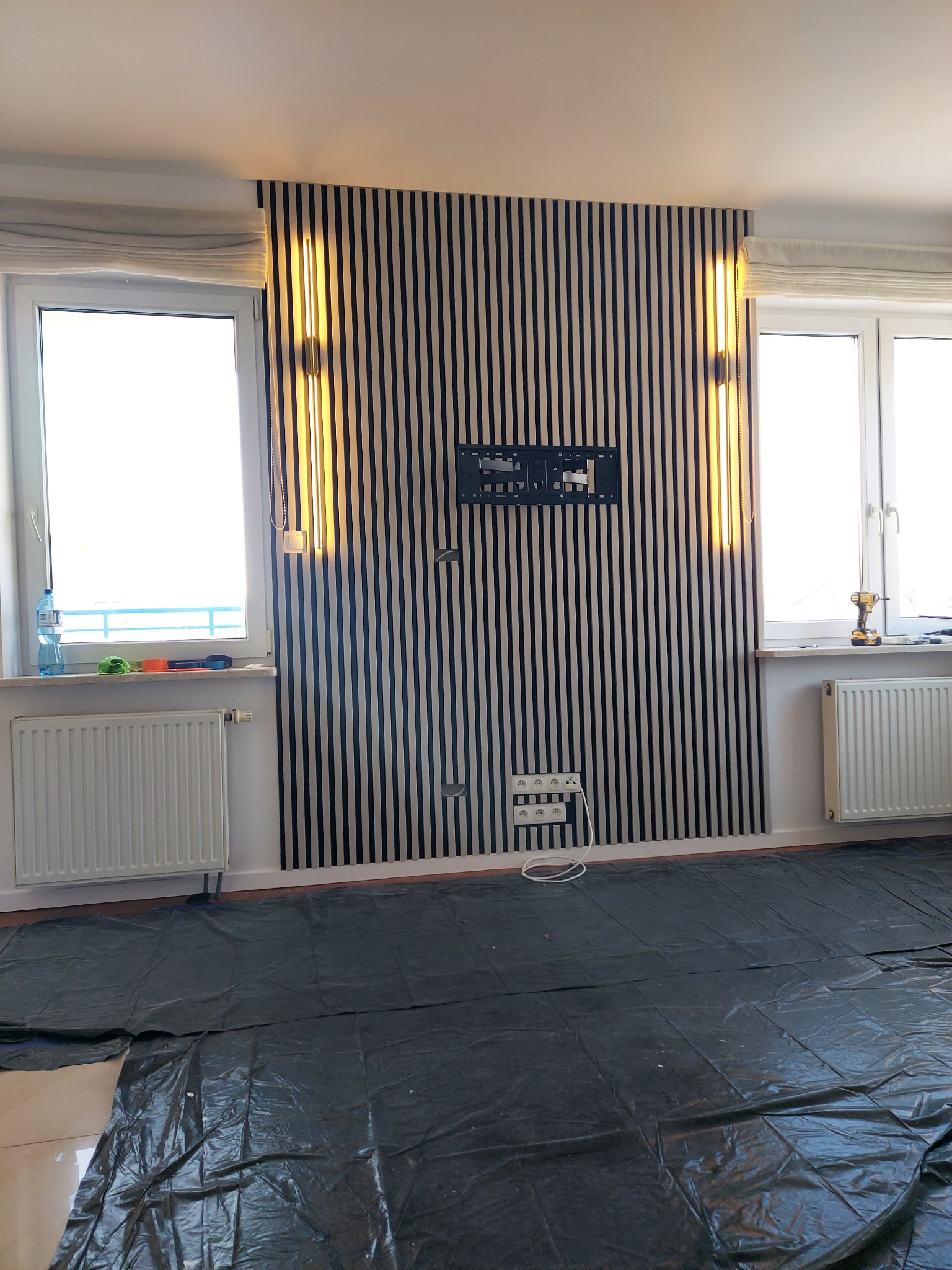 Wykończona ściana z pionowych lameli w czarno-białe pasy, z zamontowanym uchwytem na TV i lampami po bokach. Podłoga zabezpieczona folią malarską.