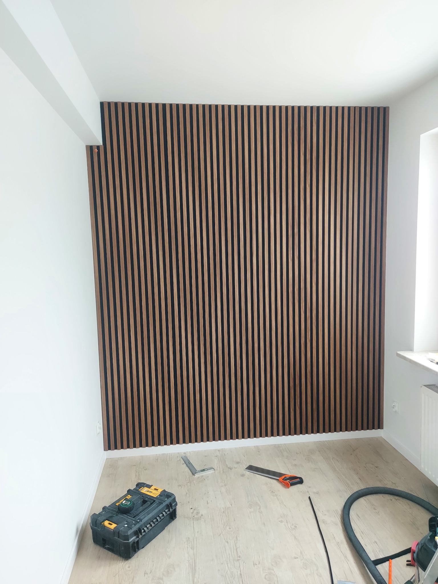 Ściana wykończona panelami lamelowymi w odcieniach brązu, narzędzia pracy na podłodze, białe ściany. Dekoracyjne wykończenie wnętrza.