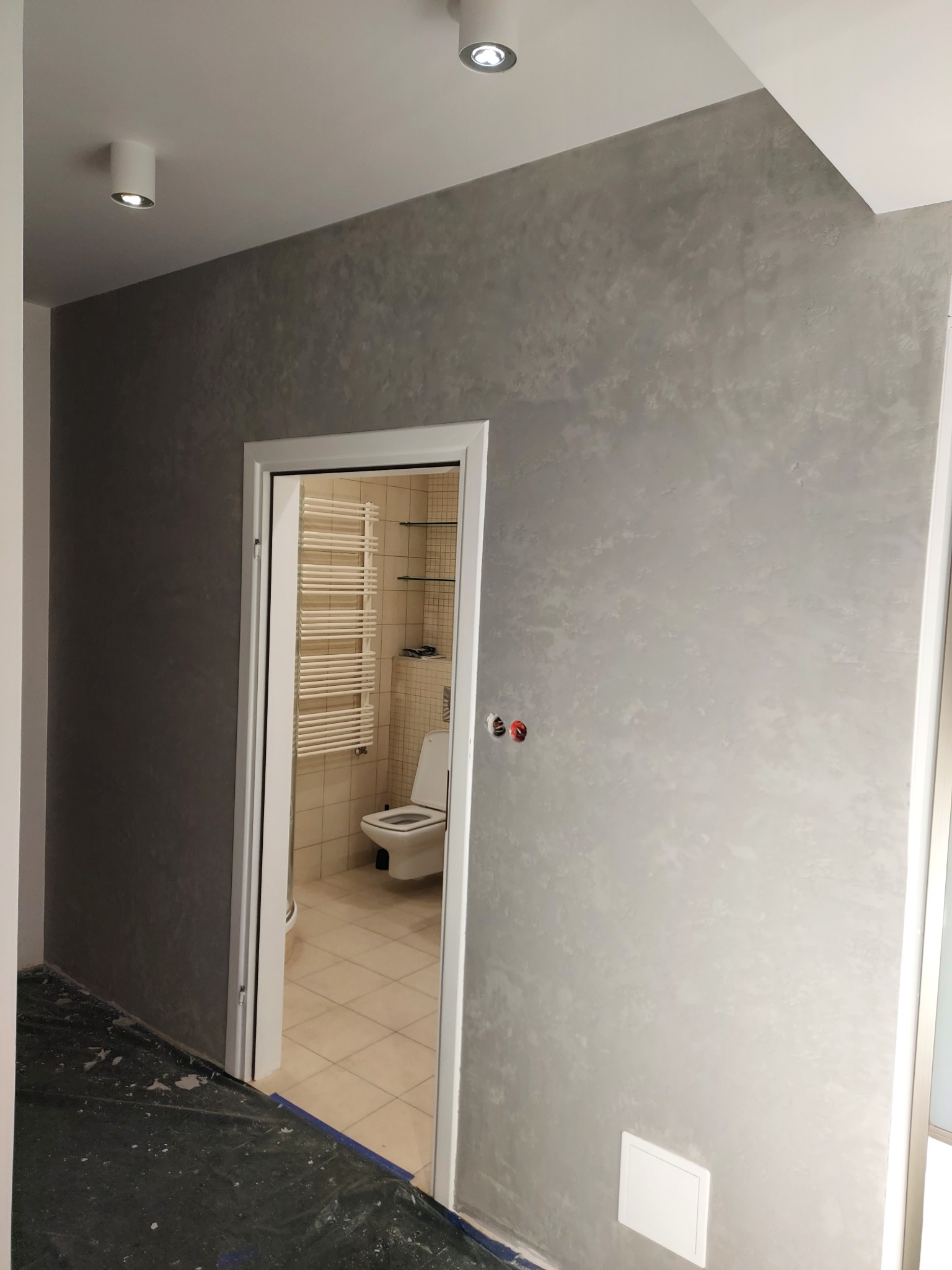 Ściana w łazience po nałożeniu dekoracyjnej, szarej gładzi weneckiej. Widoczne gniazdka elektryczne i fragment łazienki z grzejnikiem drabinkowym przez otwarte drzwi.