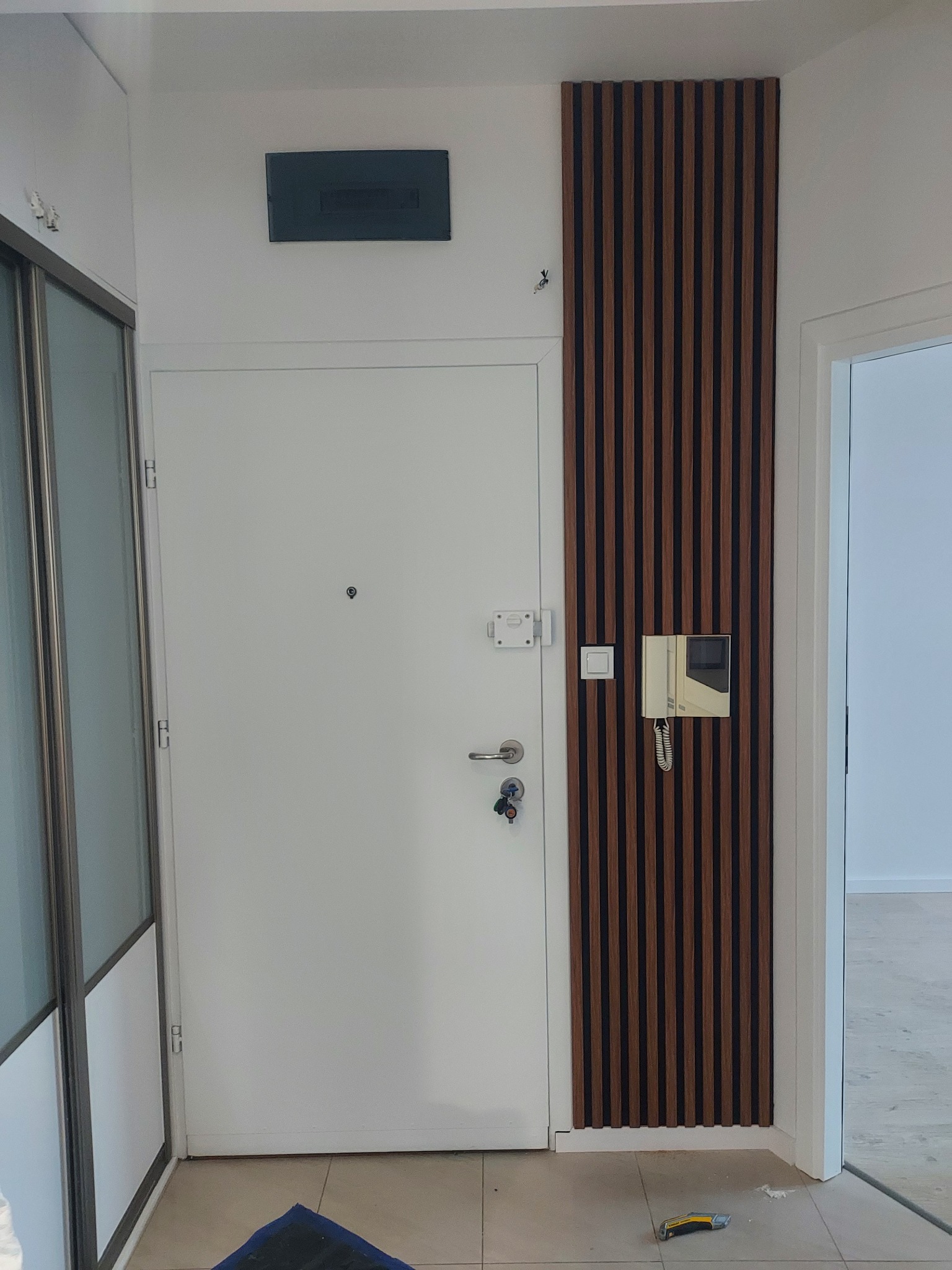 Nowoczesny przedpokój z białą szafą, drzwiami i dekoracyjną ścianką z drewnianych lameli. Widoczny domofon i narzędzie na podłodze.