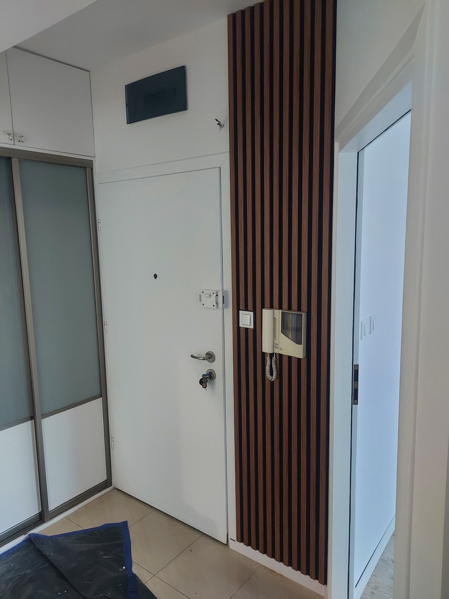 Białe drzwi wejściowe z brązowymi lamelami dekoracyjnymi, obok szafa przesuwna i drzwi wewnętrzne. Widoczny domofon i fragment podłogi z plandeką.