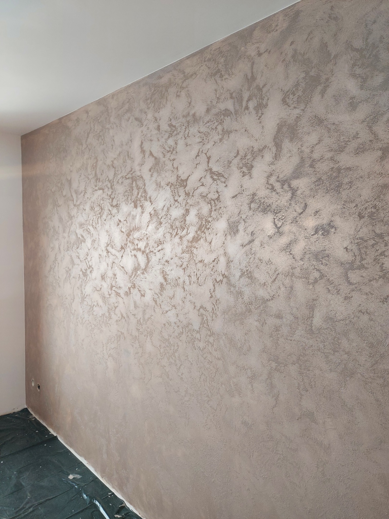 Ściana w pokoju po nałożeniu dekoracyjnej, beżowo-srebrnej gładzi. Widoczne zabezpieczenie podłogi folią malarską i fragment ściany bocznej. Efektowna faktura na ścianie.