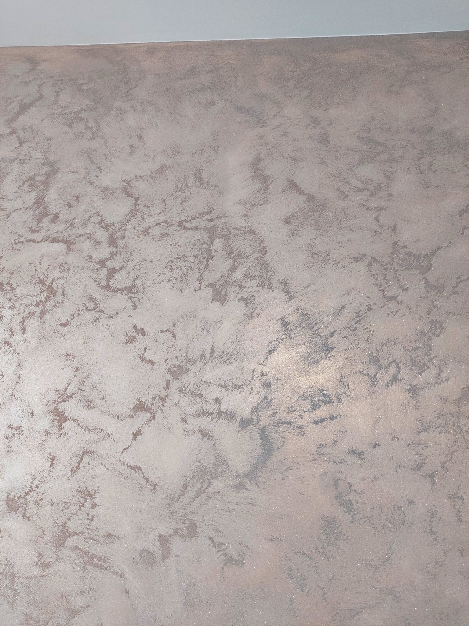 Dekoracyjna ściana z efektem metalicznego połysku i nieregularną fakturą, wykonana w odcieniach szarości i srebra, z widocznym fragmentem białego sufitu.