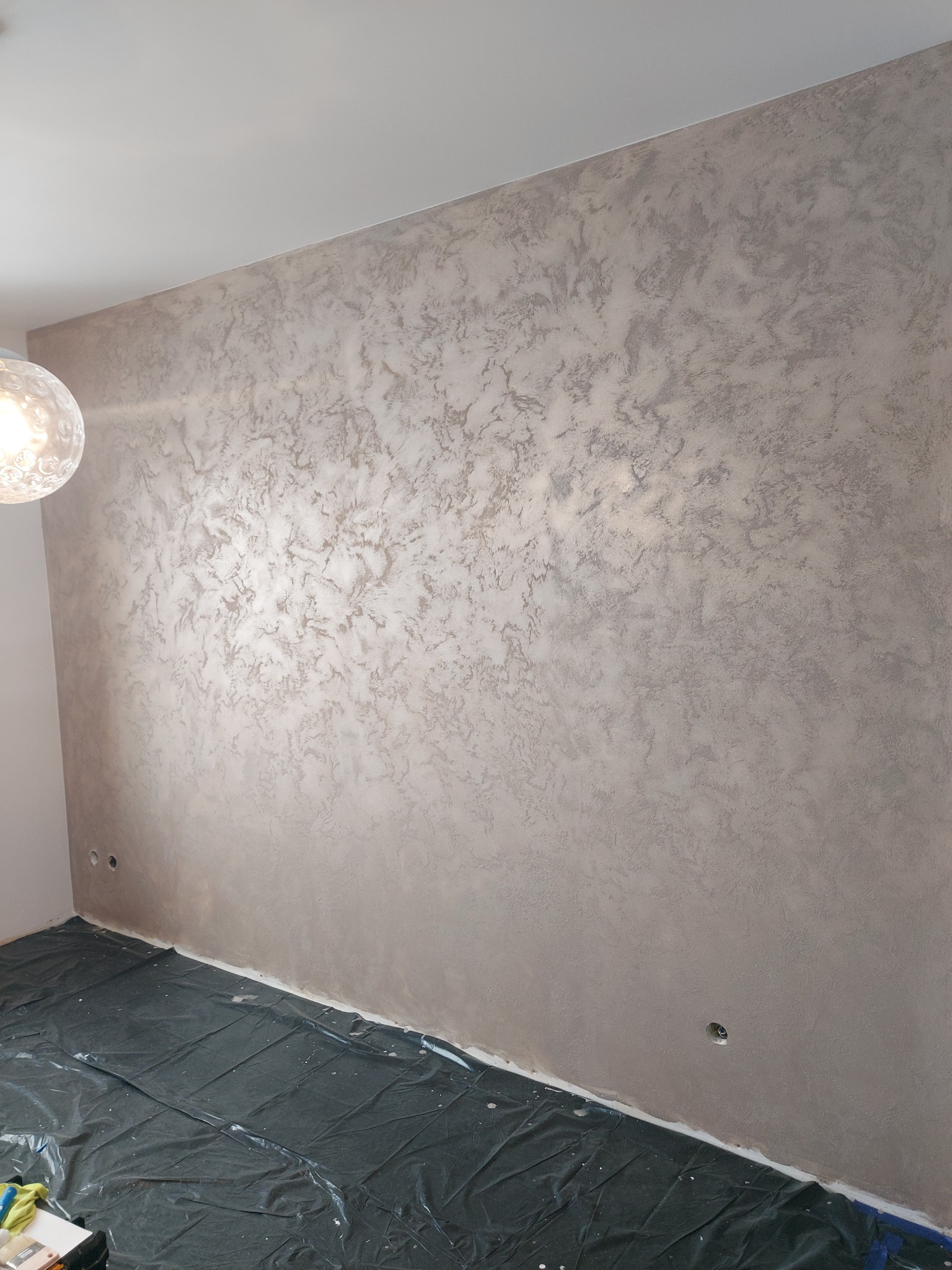 Ściana z dekoracyjną, srebrzystą strukturą, zabezpieczona folią podłoga, widoczna lampa. Efektowna faktura na ścianie w wykończonym wnętrzu.