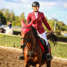 Kobieta w czerwonym żakiecie i kasku na koniu z czerwonymi ozdobami na głowie, na piaszczystym terenie z zielonym tłem i innymi jeźdźcami w oddali.