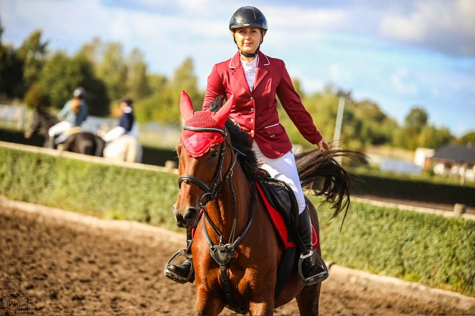 Kobieta w czerwonym żakiecie i kasku na koniu z czerwonymi ozdobami na głowie, na piaszczystym terenie z zielonym tłem i innymi jeźdźcami w oddali.
