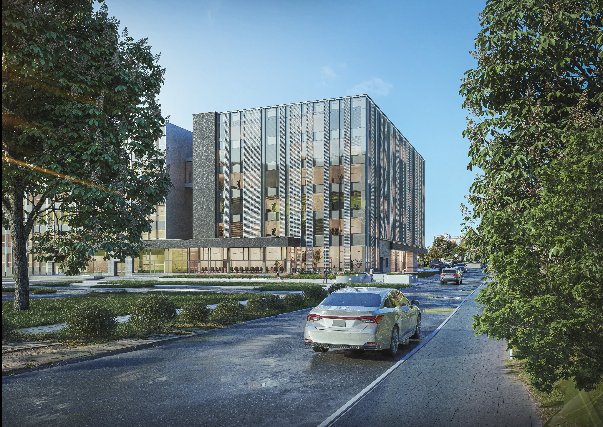 Nowoczesny budynek biurowy z fasadą ze szkła i metalu, otoczony zielenią i ulicą z samochodami. Ujęcie z poziomu ulicy, w słoneczny dzień. Widoczny fragment parkingu.