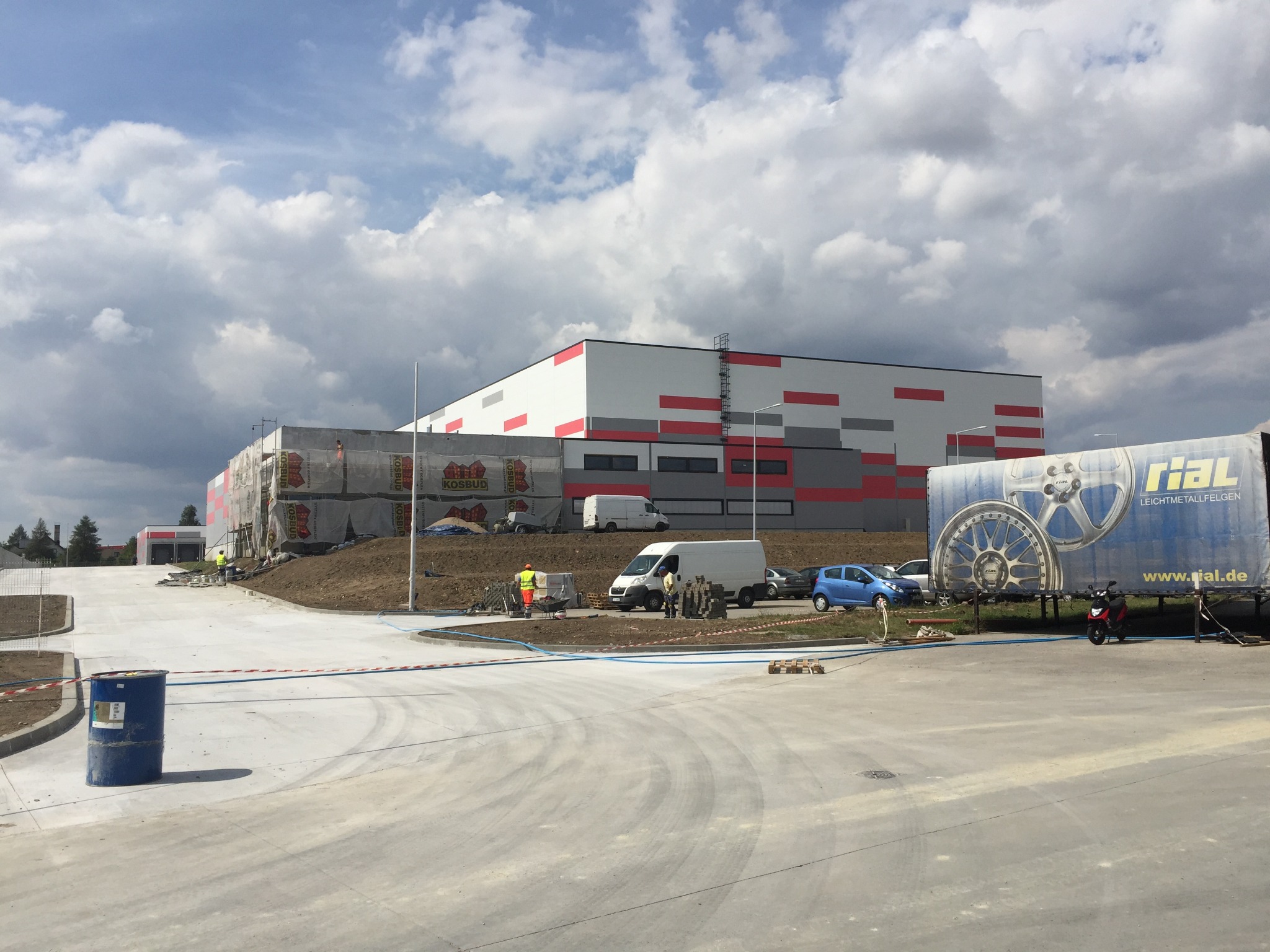 Budynek usługowy w trakcie budowy z elementami elewacji w kolorach czerwonym i szarym. Na pierwszym planie plac budowy z pojazdami i pracownikami.