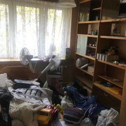 Kacper chilimoniuk - Zagracony pokój z oknem, na parapecie dwa wentylatory. Na podłodze i łóżku porozrzucane ubrania, zabawki i inne przedmioty. Regał z półkami częściowo widoczny po prawej stronie.