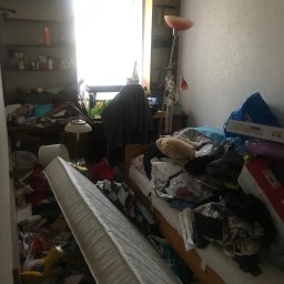 Kacper chilimoniuk - Pokój zagracony przedmiotami, z materacem opartym o ścianę, sugerujący potrzebę opróżnienia mieszkania. Widoczne półki z książkami i lampa.